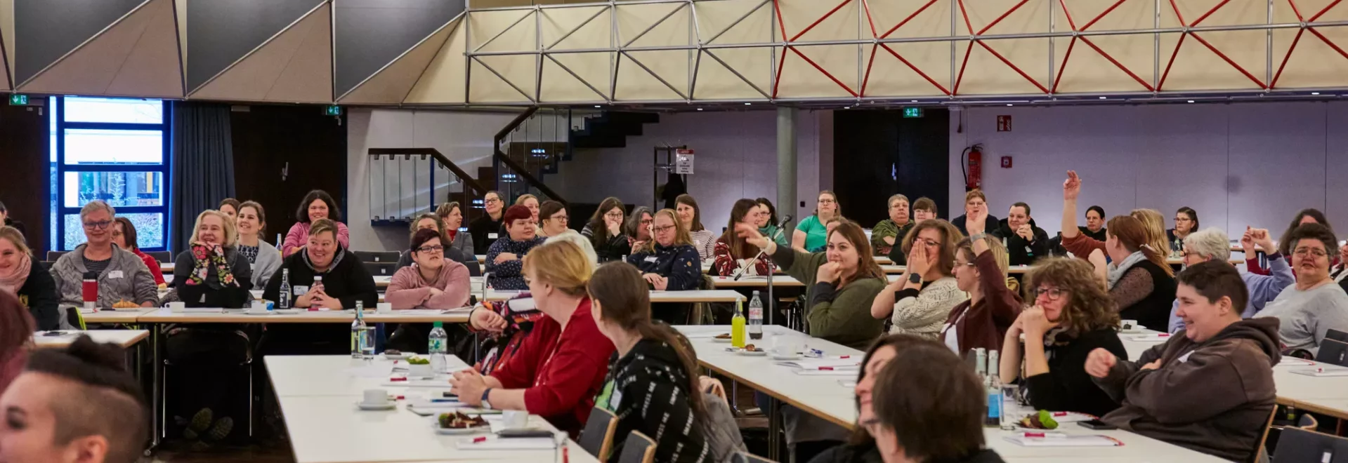 Das Bild zeigt einen großen Konferenzraum, der voller Frauen ist. Viele Personen sitzen an langen Tischen und schauen nach vorne. Einige Personen lächeln, andere scheinen aufmerksam zuzuhören oder miteinander zu sprechen. Die Tische sind mit Wasserflaschen, Notizbüchern und anderen Materialien bedeckt. Im Hintergrund sieht man eine erhöhte Galerie und moderne Wandelemente. Die Atmosphäre wirkt lebendig und konzentriert