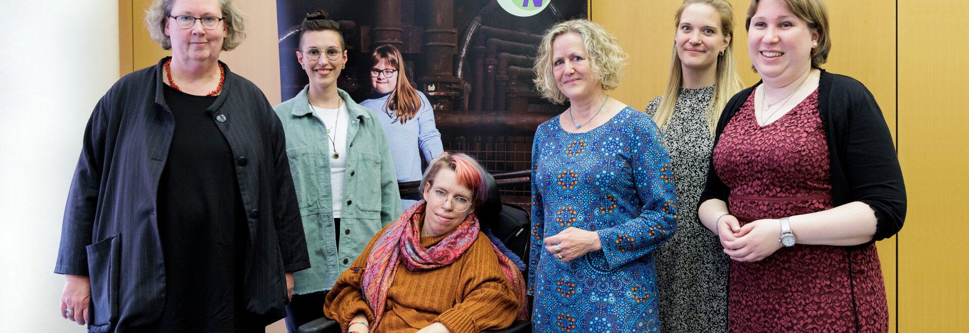 Auf dem Bild sind die sechs Frauen zu sehen, die zusammen im NetzwerkBüro arbeiten. Eine der Frauen sitzt in einem Rollstuhl. Sie tragen verschiedene Outfits, von lässig bis elegant. Im Hintergrund ist ein Poster mit dem Logo vom Netzwerk NRW und dem Bild eines Mädchens mit Down-Syndrom zu sehen. Alle Frauen lächeln freundlich in die Kamera und die Atmosphäre wirkt professionell und herzlich